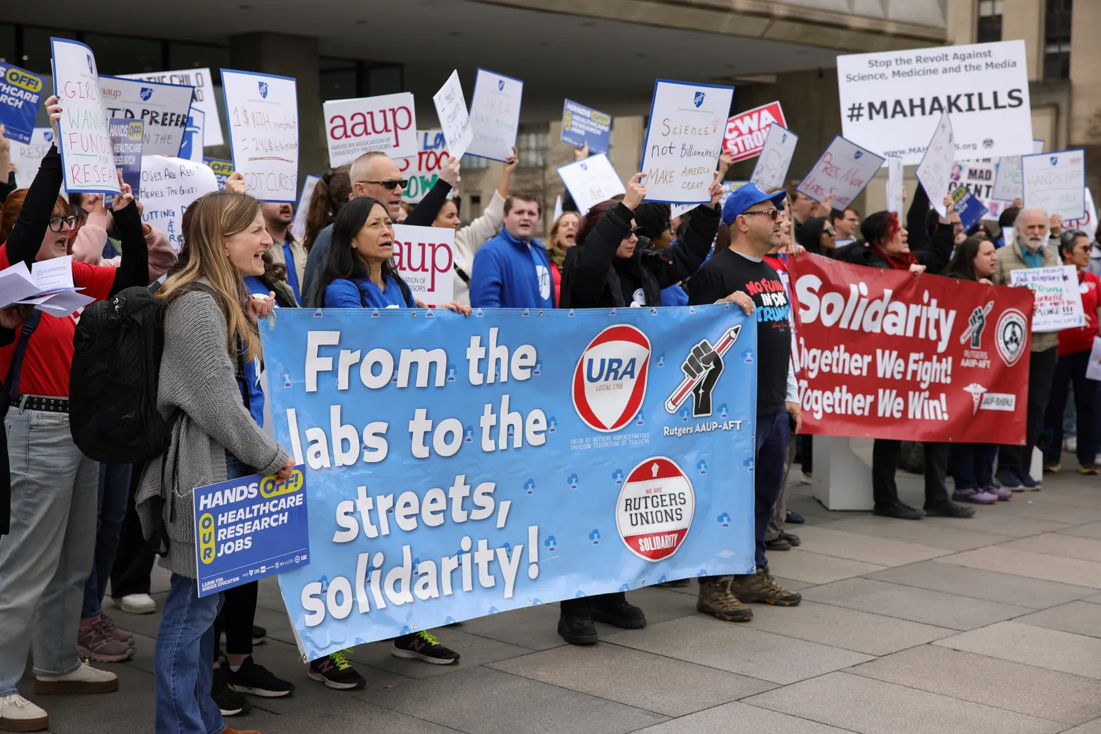 A crowd of people hold signs in solidarity with HHS employees. A banner reads, “From the labs to the streets, solidarity! and “Rutgers Union”.