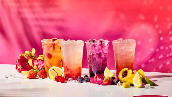 Four plastic cups with colorful beverages and ice inside sit on a white table, surrounded by fruit and against a pink patterned background.