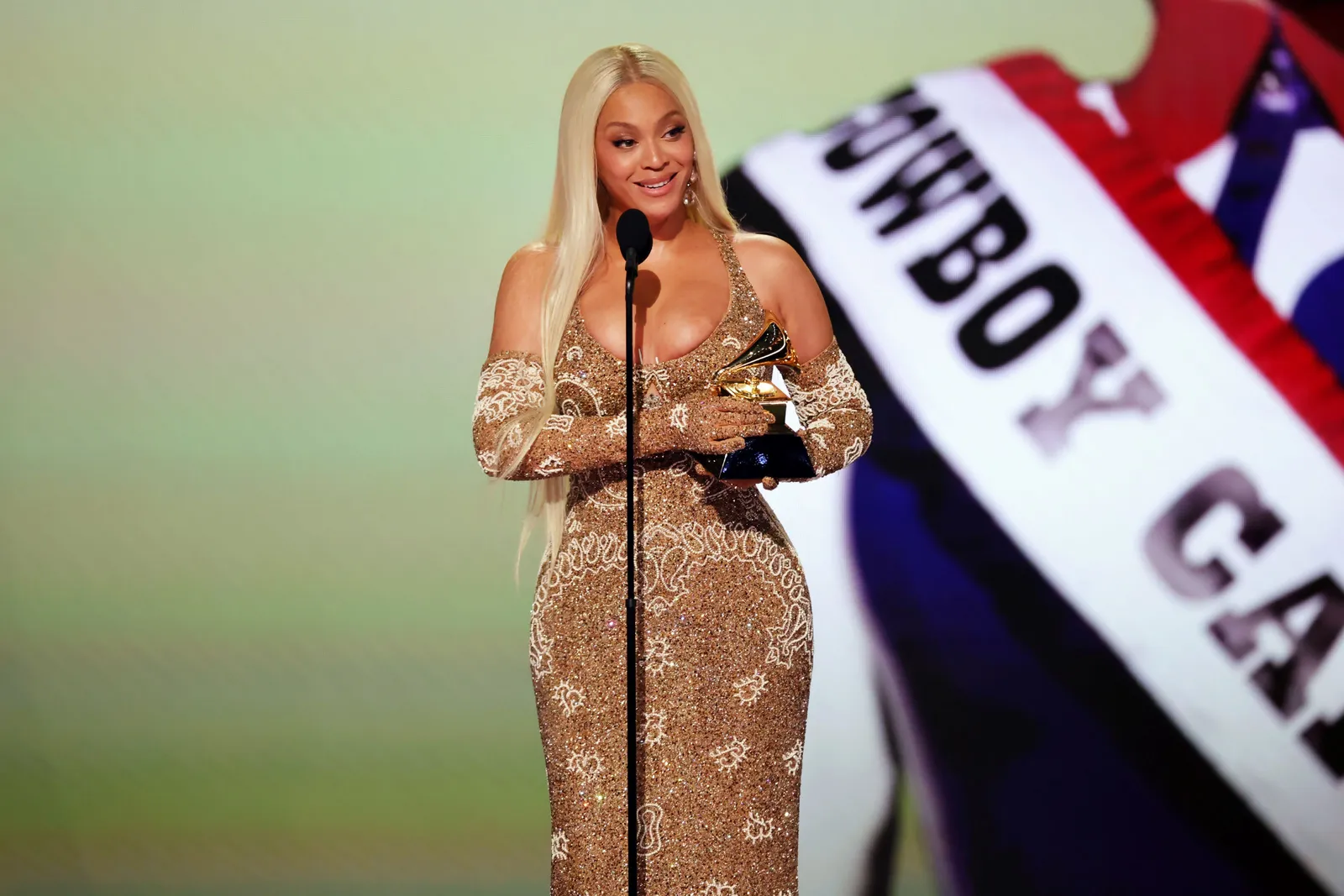 A person in a gold dress and opera gloves stands on a stage holding a Grammy award.