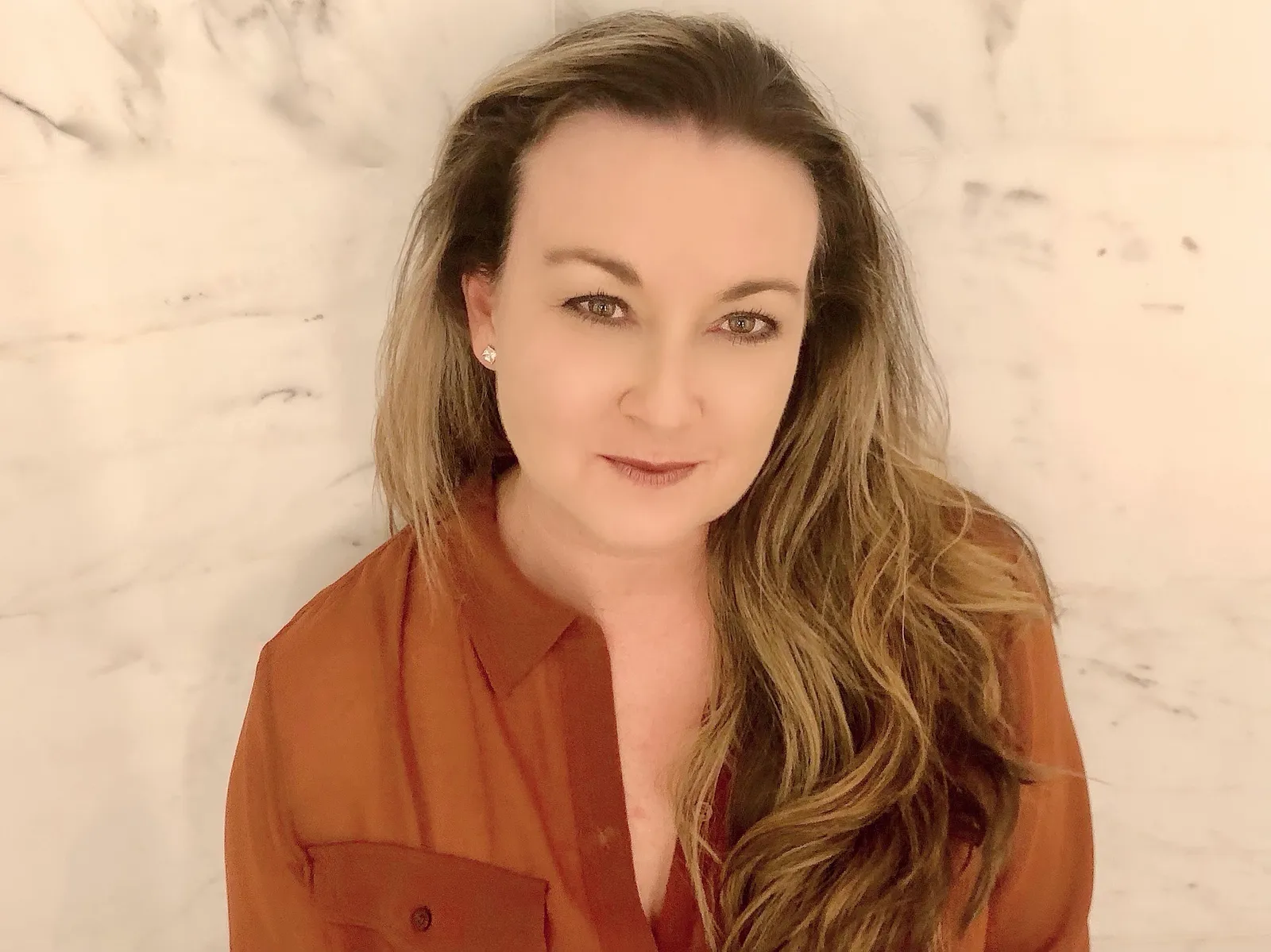 A corporate headshot shows a woman in a sheer brown blouse and long blonde hair leaning against a marble wall.