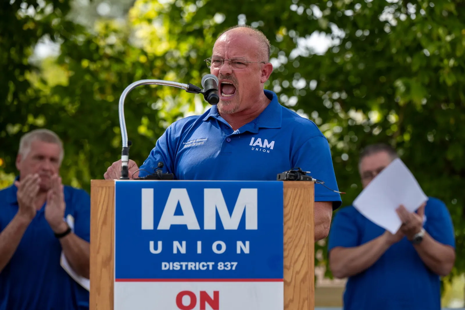 A man in a blue polo shirt standing behind a podium and yelling into a microphone.