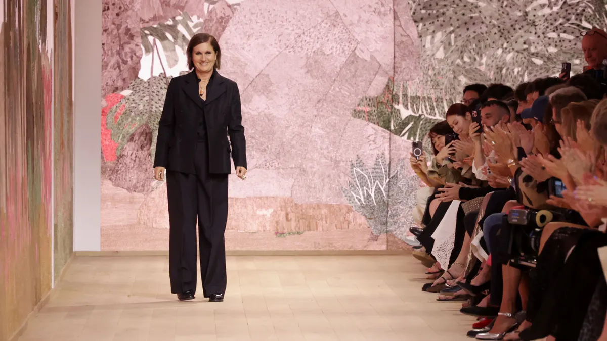 Maria Grazia Chiuri stands on the runway while a crowd applauds her.
