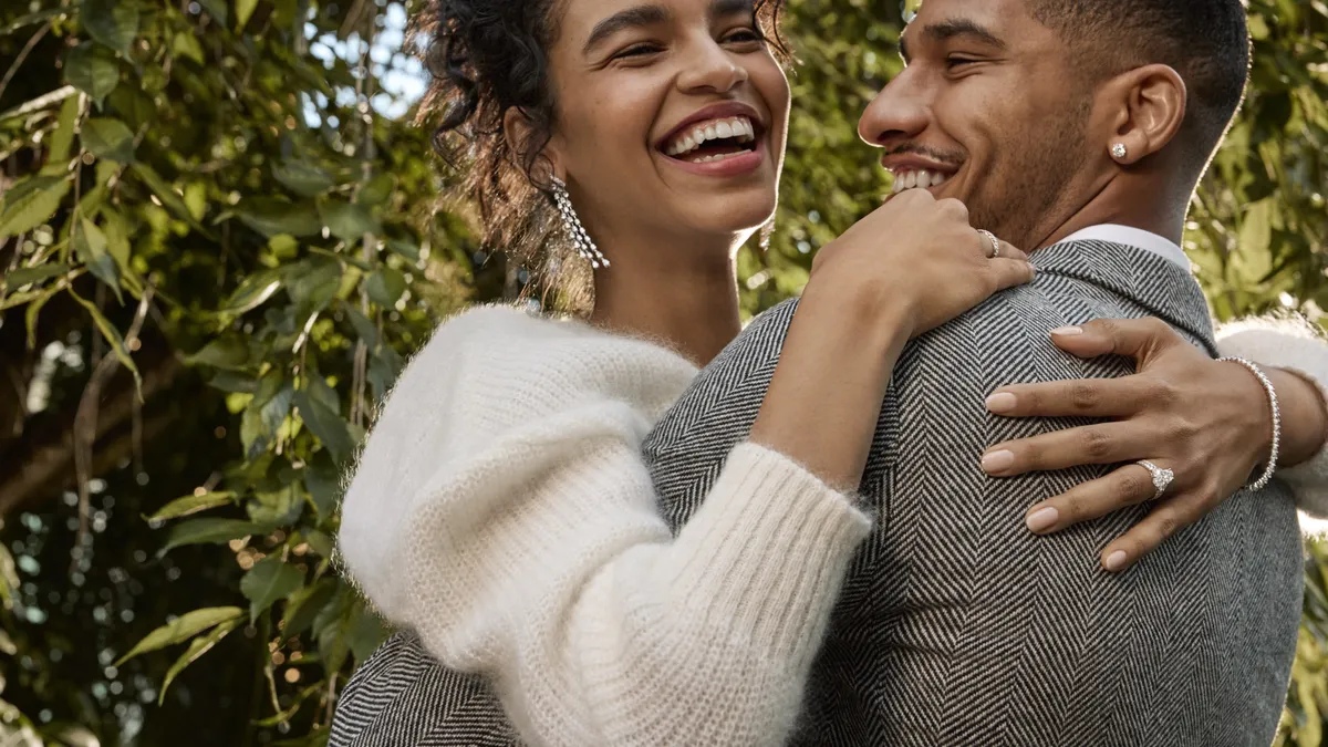 A couple wearing several pieces of jewelery embraces outdoors in the daytime.