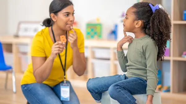 An adult is kneeling on the ground in a classroom. They have a finger to their lips and holding the letter B. A student is sitting across from the adult on a bench and has a finger to their lips