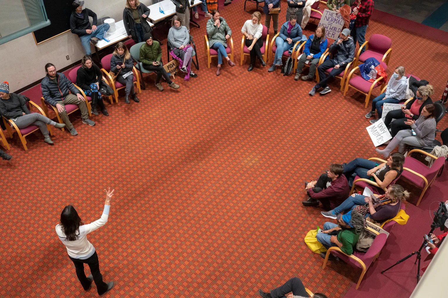 An overhead view shows people sitting in chairs in a U-shape inside a room. One person is standing in front of the people.