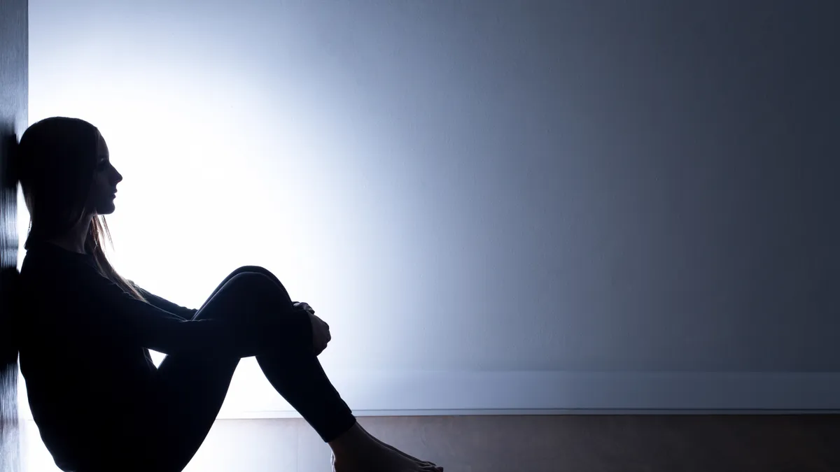 A side view of a teen sitting on the floor with back against the wall in dark hallway with a light in the background