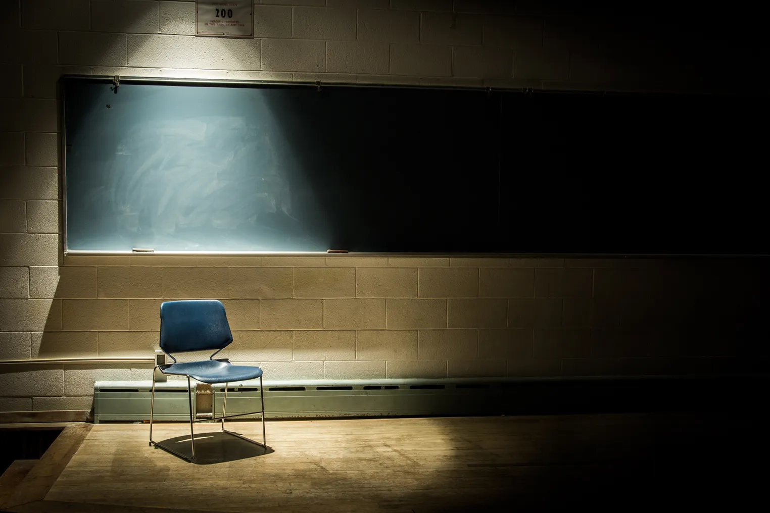 An empty chair sits in the front of a classroom. Behind the chair is a blackboard and the chair has a spotlight over it in the dark room.