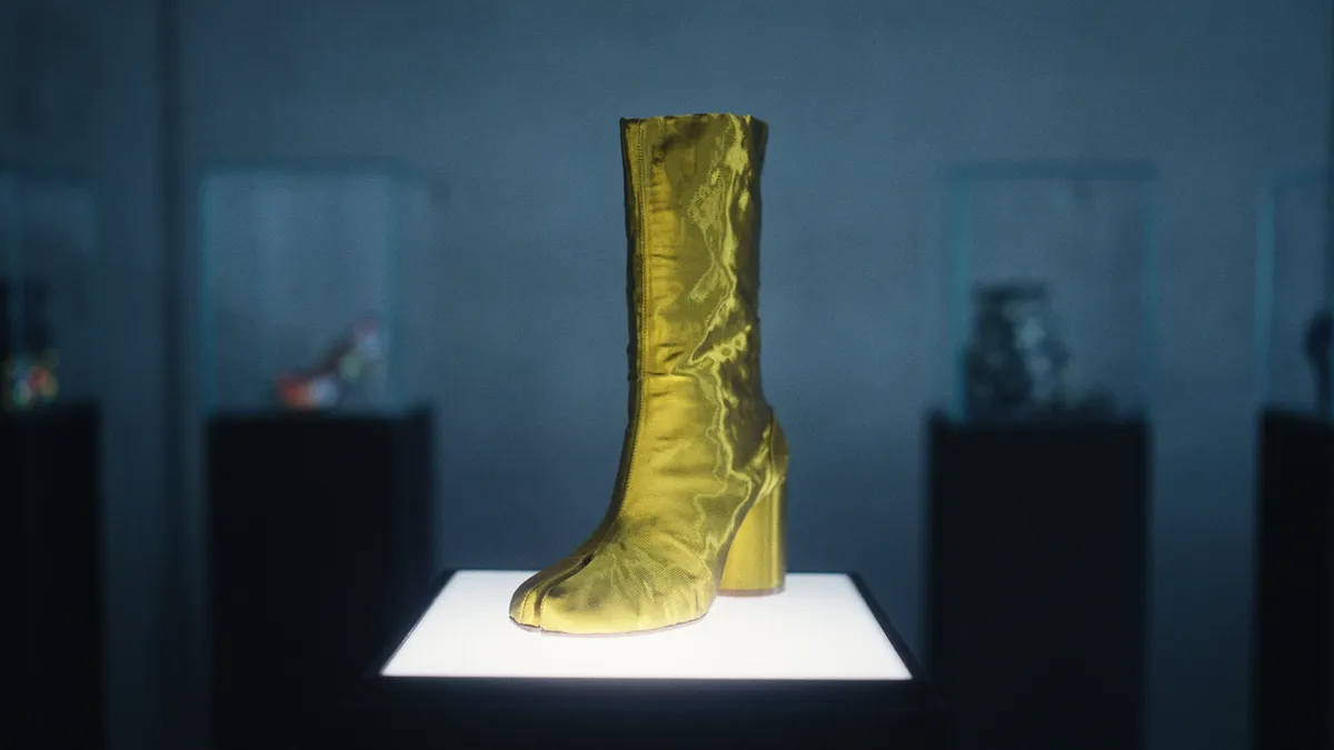 A gold Tabi split toe boot sits on a clear stand.