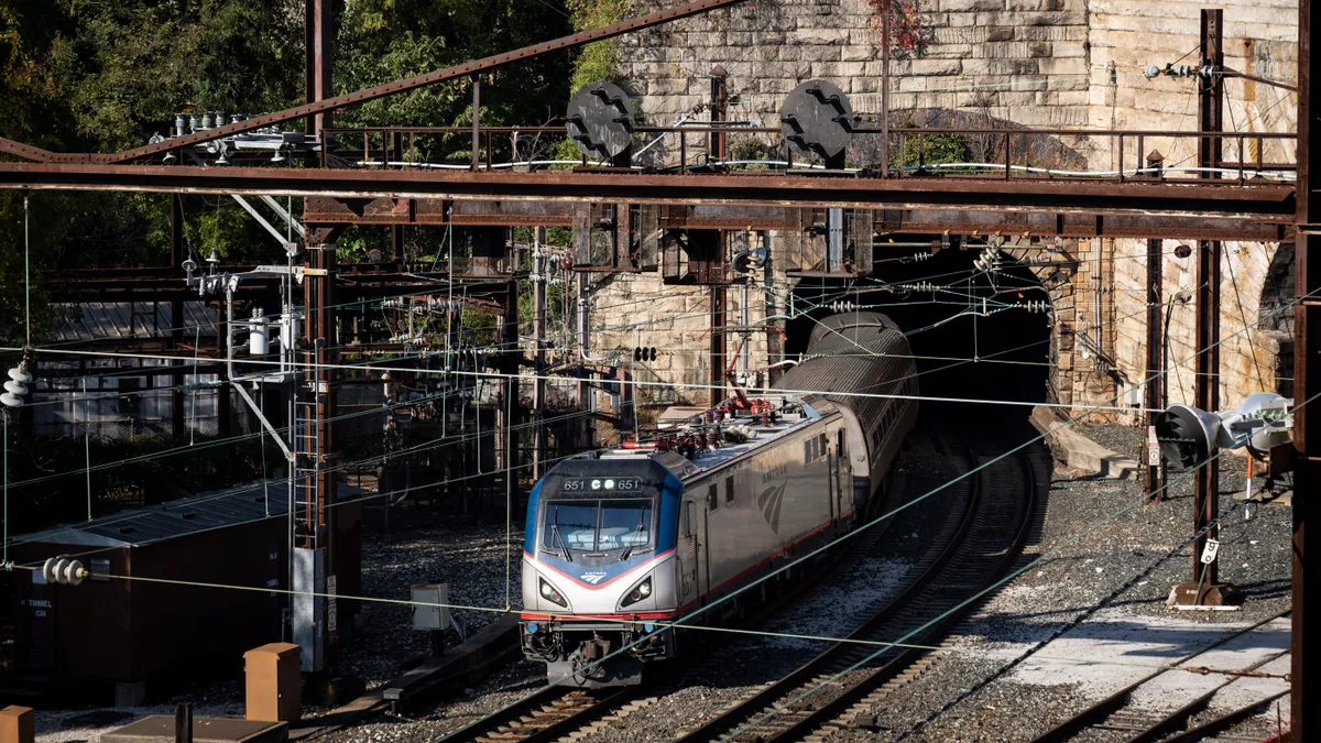 A silver Amtrak train comes out of the opening of a brown stone tunnel.