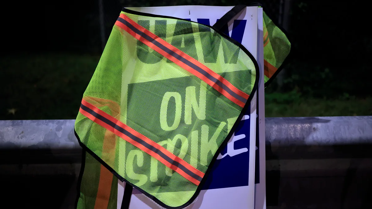 A UAW picket sign is pictured, with a neon yellow mesh vest (with neon orange stripes and light-reflective strips) tossed over it