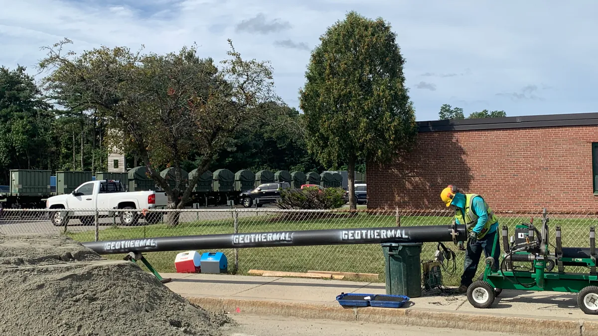 A worker in a hard hat and yellow vest lays a large pipe in the ground. On the side of the pipe, the word "geothermal" is written three times.