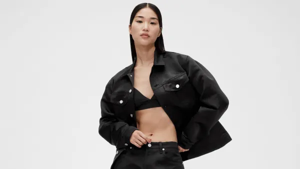 A model wears a black satin cropped jacket over a bikini top and loose fitting pants and stands against a white backdrop.