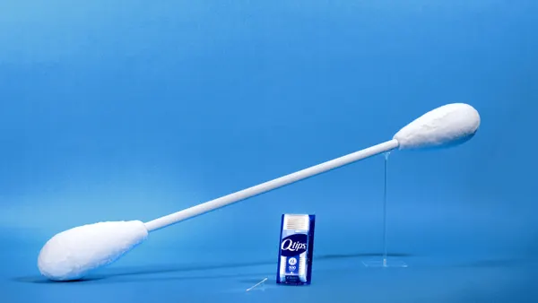 A giant Q-tip stands above a standard package of the cotton swabs against a blue background
