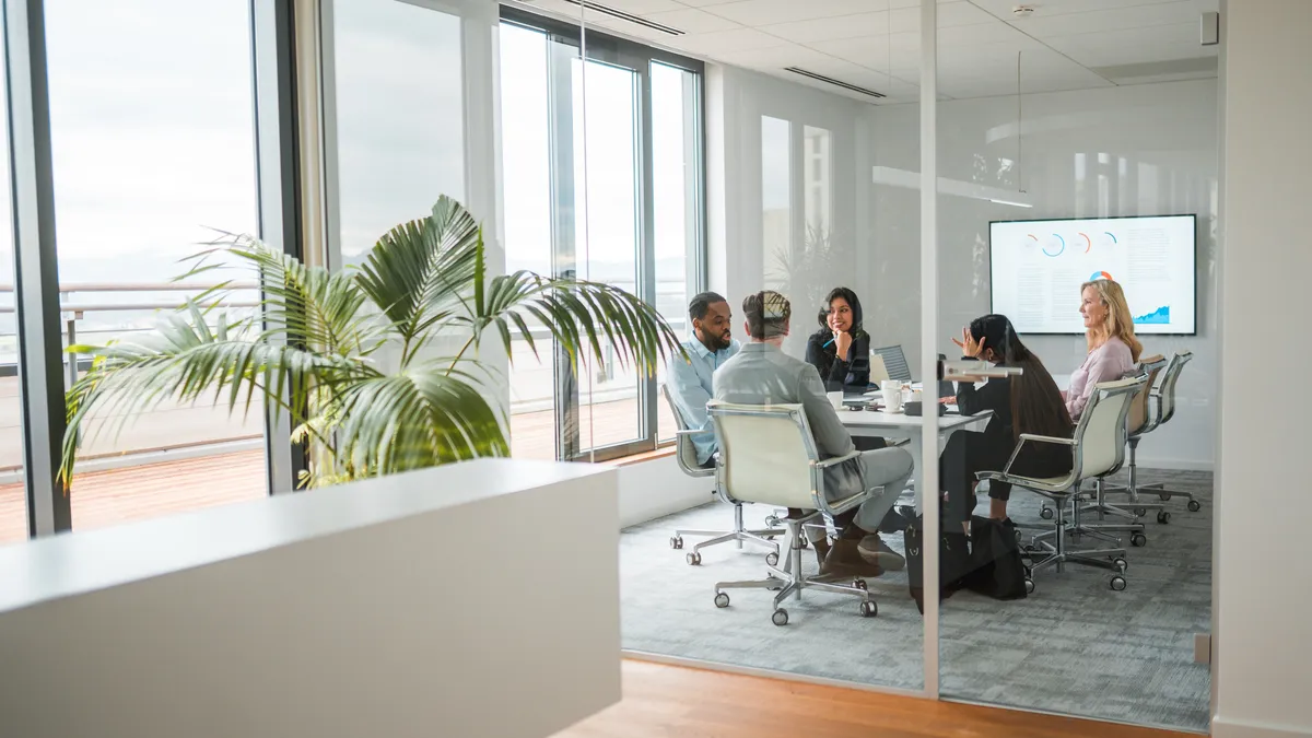 A wide angle shot of a multiracial team sitting in a closed meeting room of a modern office. Shot through glass walls, with a big potted plant next to large windows overlooking the patio.