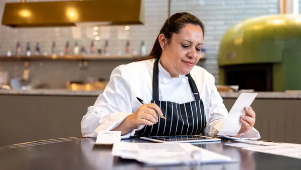 A person wearing a black apron with white stripes and white shirt sits at a metal table looking at receipts.