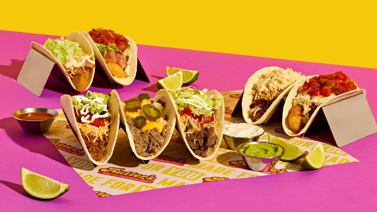 A photo of multiple tacos, each with different toppings, in taco holders and sitting on a pink surface against a yellow background.