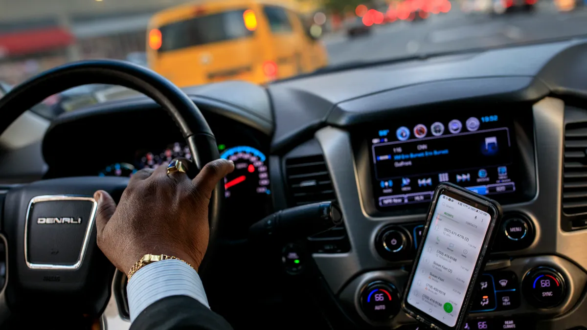 Car driver's hand on the wheel with a smartphone to the right.