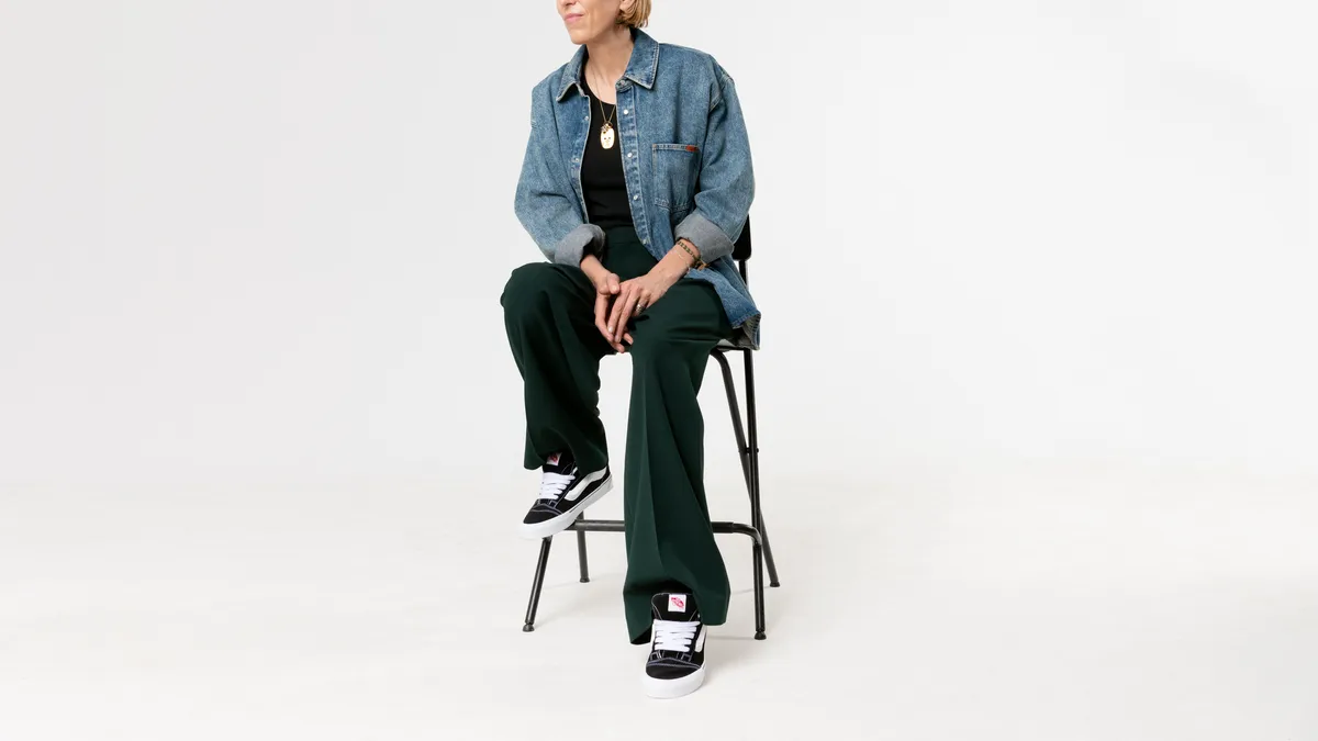 A person wearing a denim jacket, green pants and black-and-white sneakers sits on a stool.