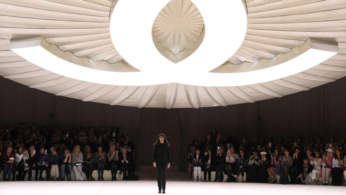 A person stands beneath a giant Chanel logo on a dark runway.