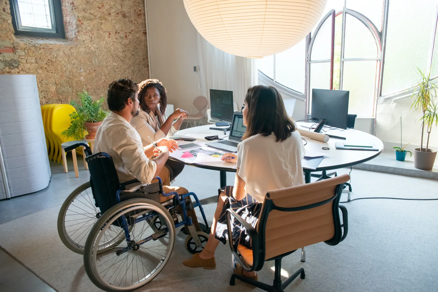 A group of coworkers sit at a table, discussing work. One of the coworkers is seated in his wheelchair.