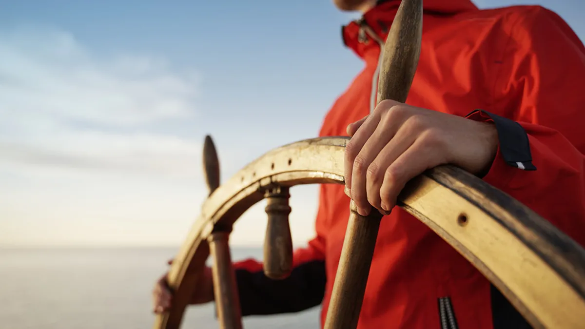 A persons hands on a wooden steering wheel looking out into the ocean