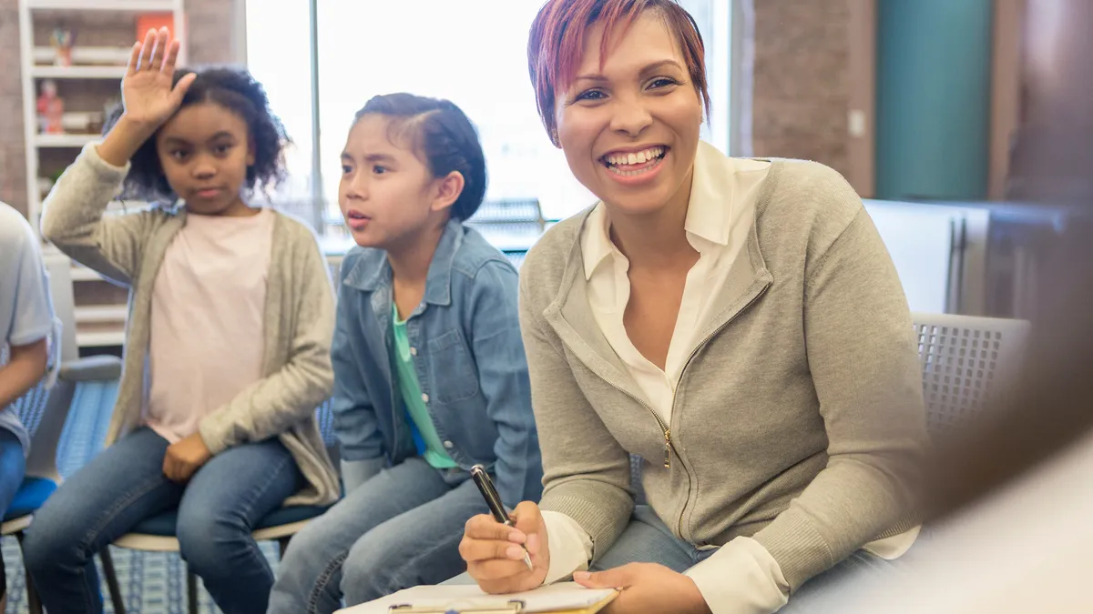 A teacher leads a classroom discussion as one student raises their hand during the conversation.