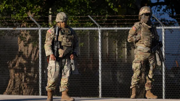 Two people dressed in military fatigues with masks over their faces and holding guns in front of barbed wire fence.