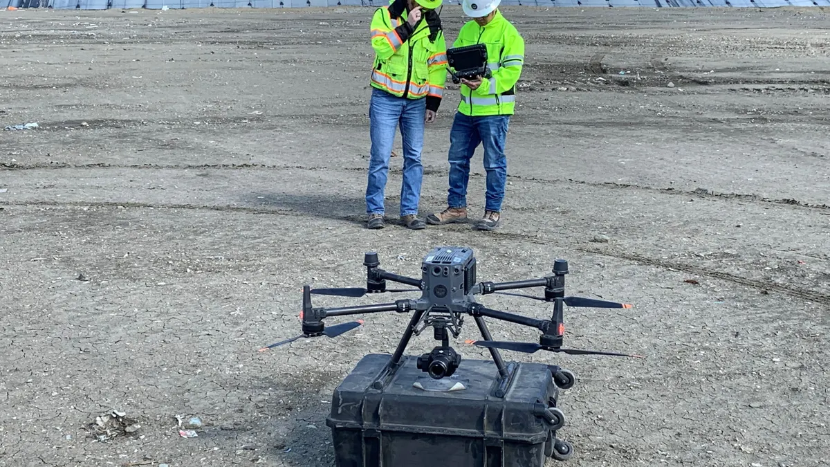 A drone rests atop a carrying case while two people wearing hard hats and construction vests consult a tablet behind it