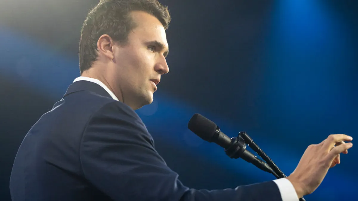 Charlie Kirk stands behind a microphone with his hand raised in gesture