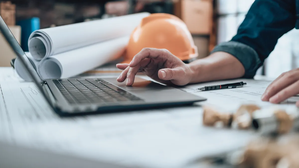 A person works on a laptop in the foreground of an artistically minded shot. In the background are blueprints and a yellow hardhat.