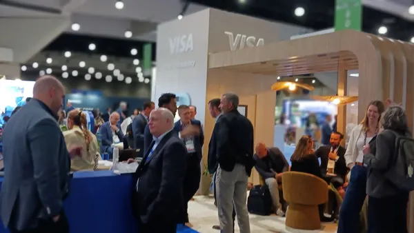 People scattered across a conference scene talking, standing, with some at high top tables and Visa booth in background.
