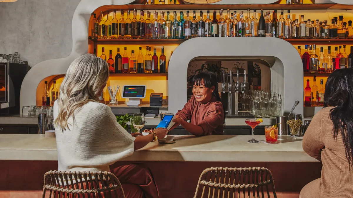 Two people are seated at a bar. A bartender uses a mobile device to scan a one of the customer's credit card while smiling.