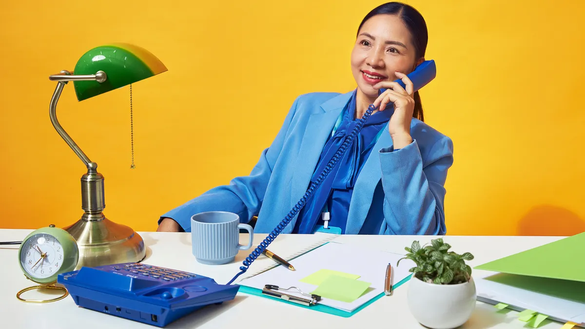 A woman in a blue suit is smiling while talking on a blue corded landline phone in a brightly colored office setup.