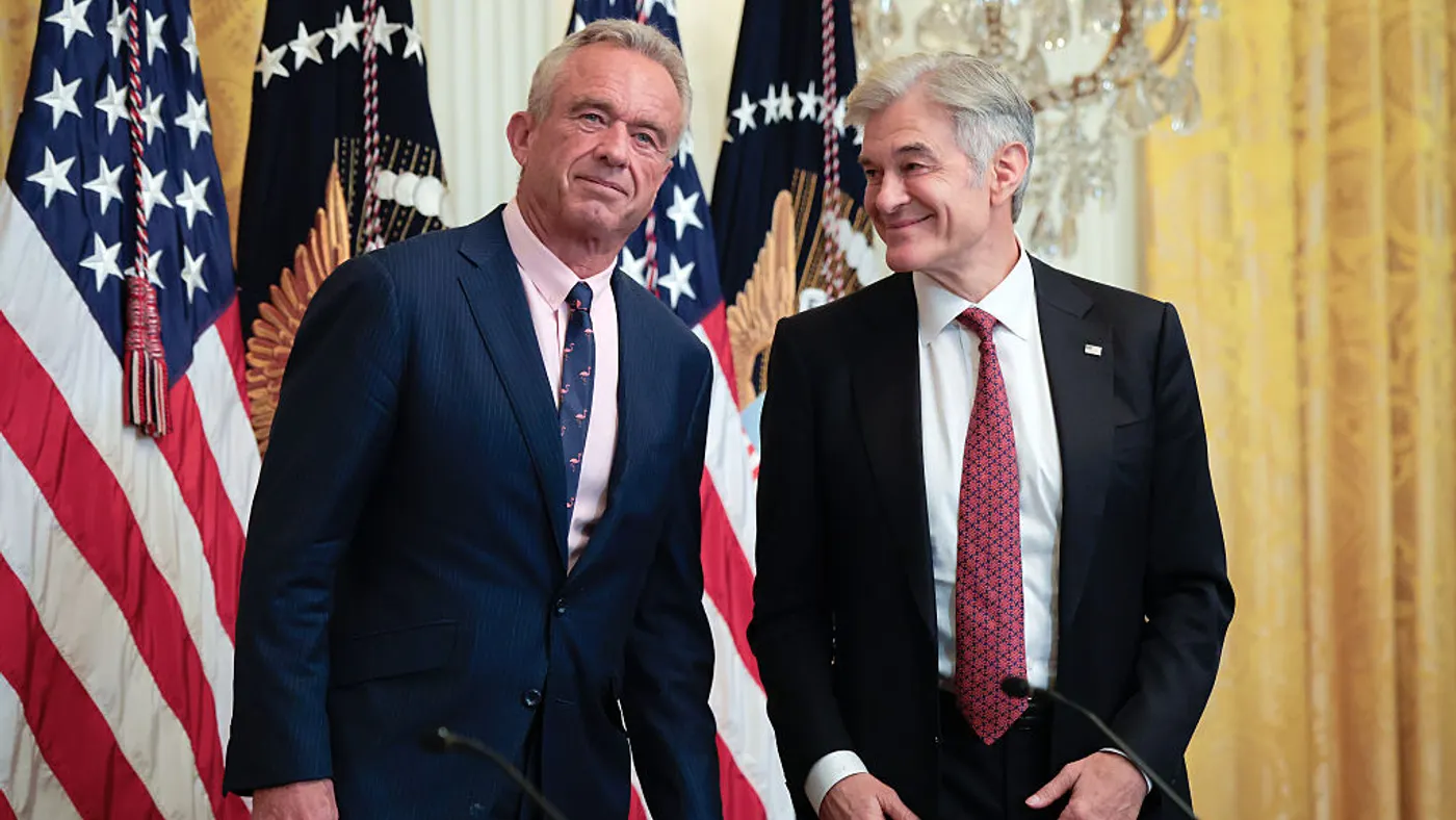 Two men in dark suits stand in front of a row of American flags.