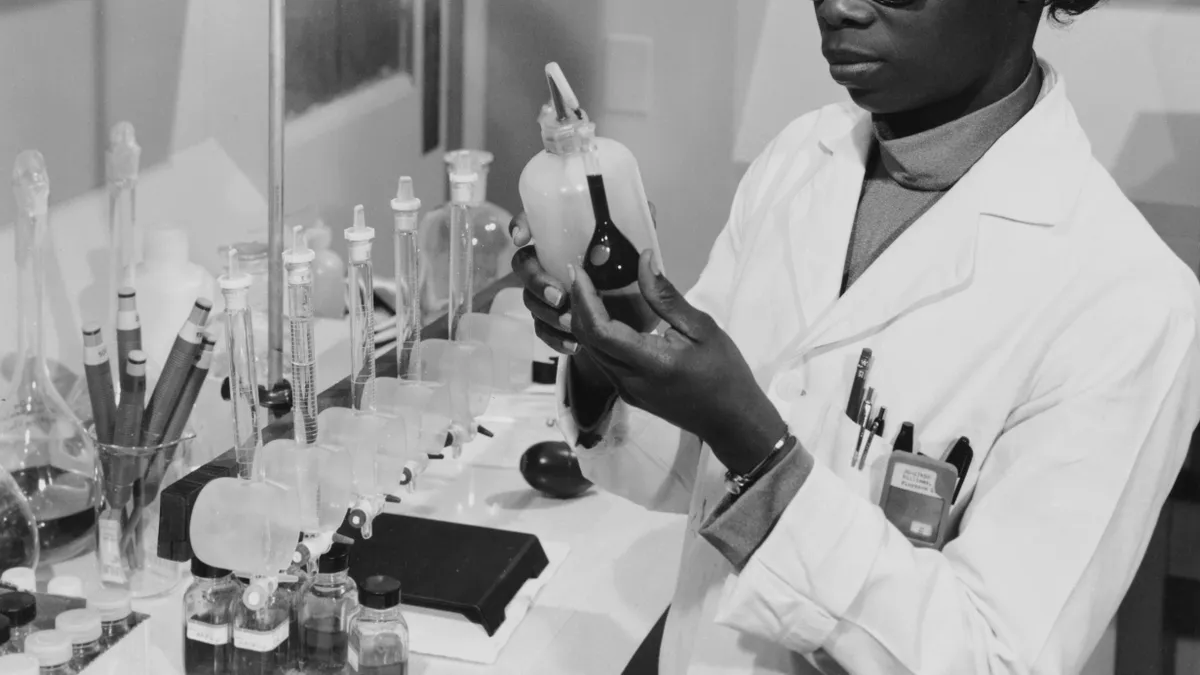 Black and white: A beautiful dark-skinned Black woman with perfectly coiffed hair sits in a lab, looking intently at a little dark glass bulb flask