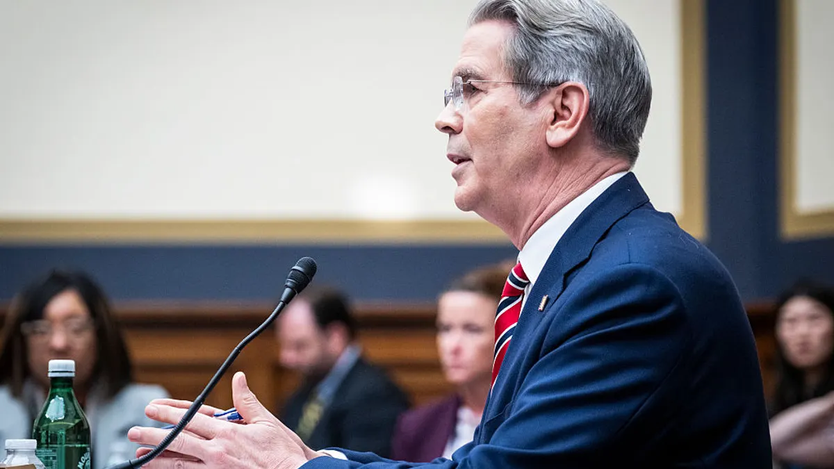 Scott Bessent, secretary of the Treasury Department, speaks into a microphone while gesturing with his hands, with blurred onlookers in the background.