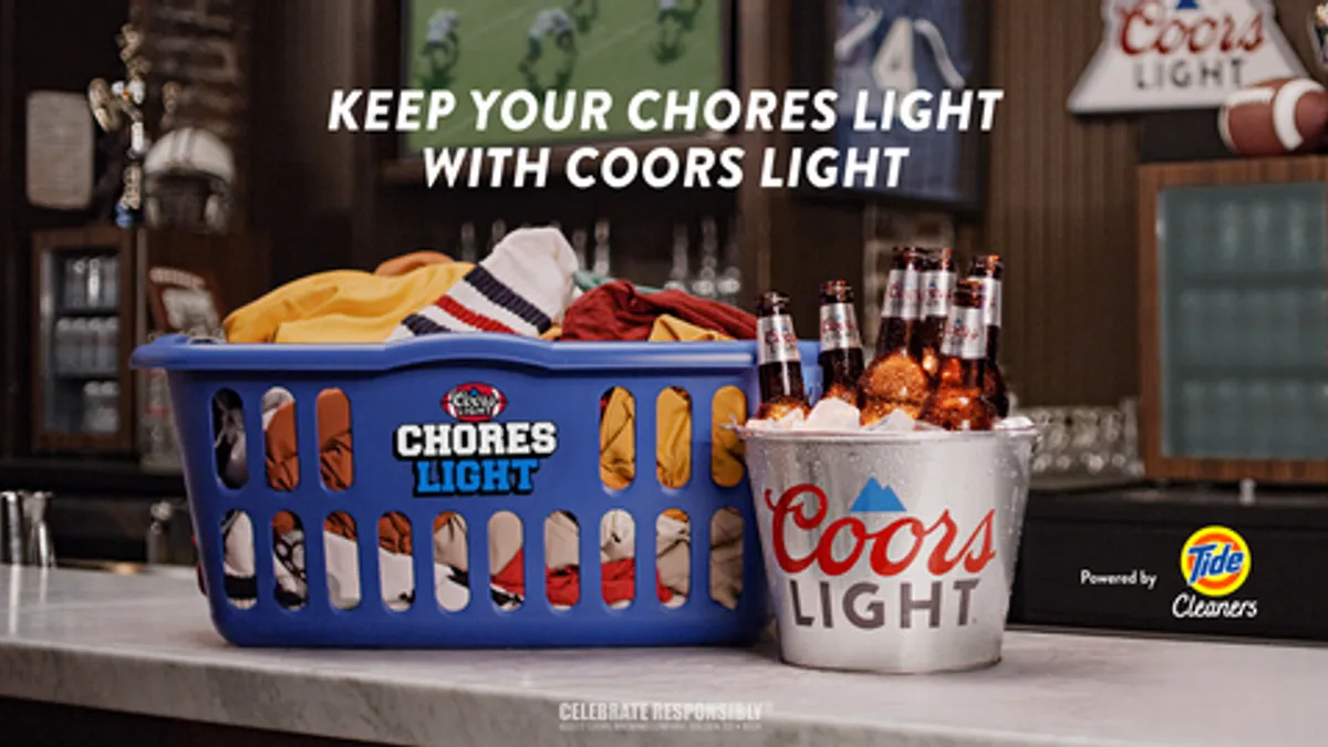 A basket of dirty laundry in a blue hamper with the text Chores Light on the front sits next to a Coors Light bucket filled with beers and ice on a bar top,