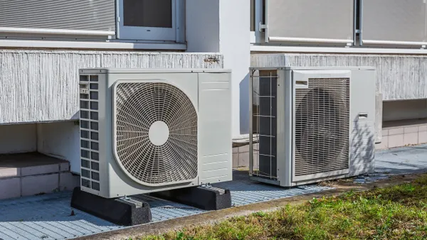 Two large outdoor HVAC units are installed on a concrete platform outside a building. Each unit has a visible circular fan and a rectangular vented casing. The building’s exterior includes gray walls, windows with closed blinds, and a strip of weathered wood paneling. Some grass and concrete blocks are in the foreground.