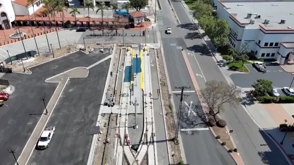 Overhead view of a street alongside two railroad tracks and a parking area with a red tile roofed train station.