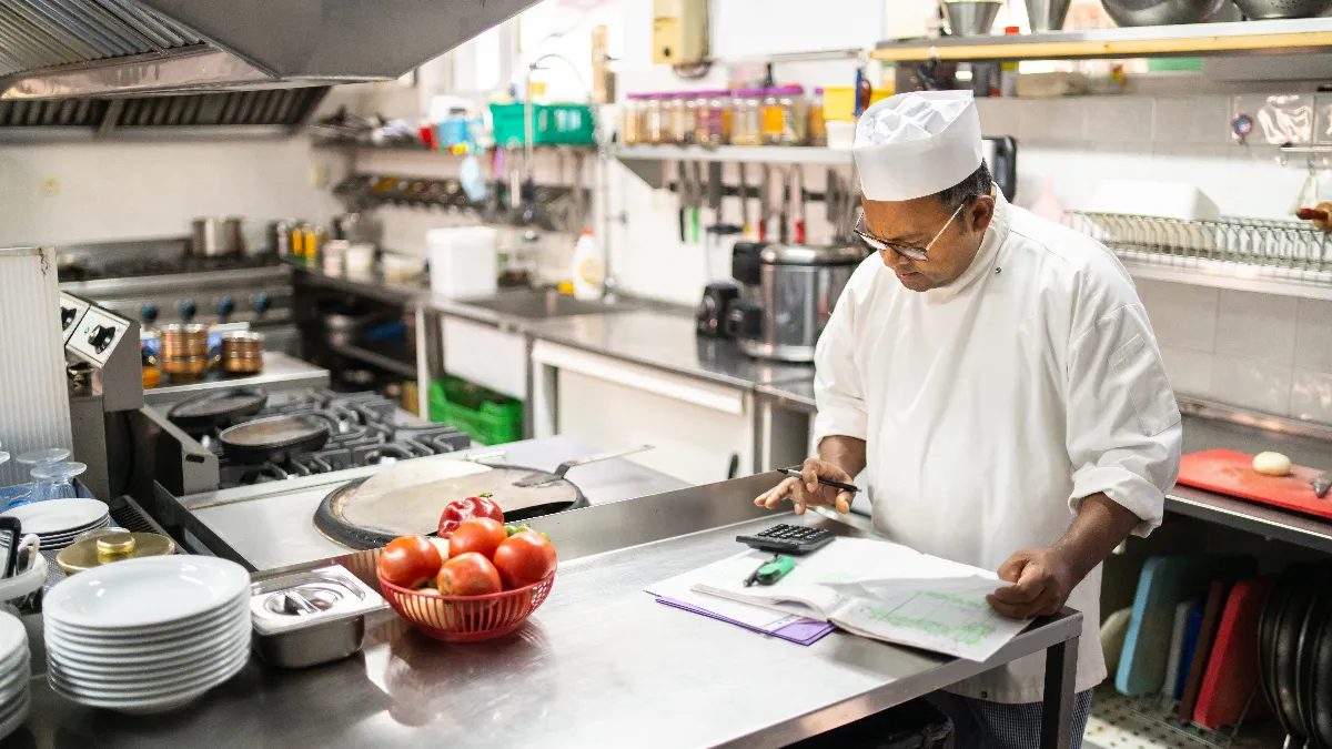 A picture of a chef in a kitchen looking down at a calculator.