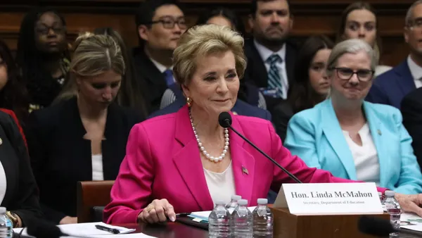 A person sits at a table with a microphone. Water bottles are nearby and people are seated in rows behind the person.