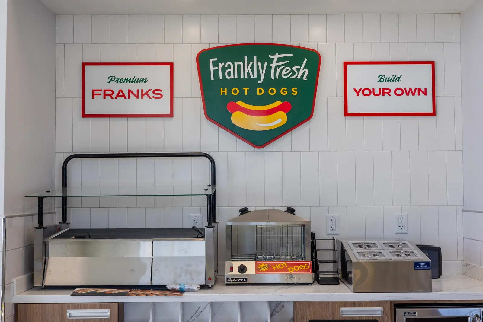 A roller grill and space for hot dog fixings sit on a counter against a white tile wall. Signs on the wall say Frankly Fresh Hot Dogs. Premium Franks. Build your own.
