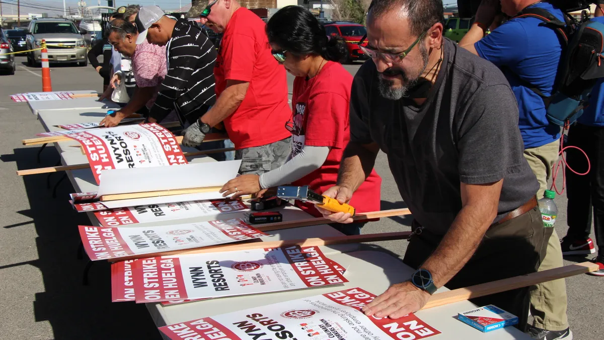 Picket signs being assembled by a group of people on plastic tables outdoors.