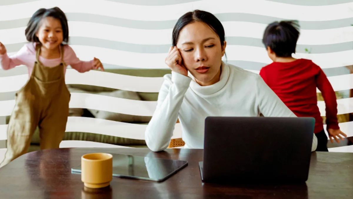 A person is looking tired at a laptop on a table while a child jumps up in the background and another looks away. There is a yellow mug on the table.