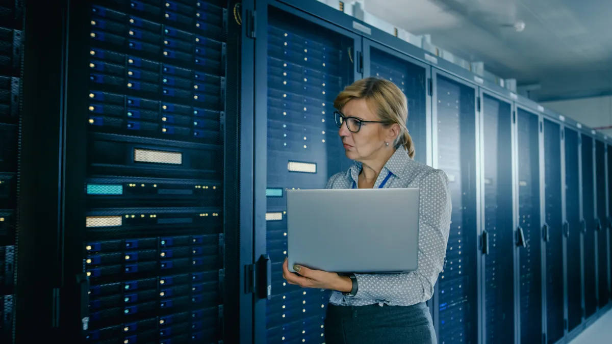 In Data Center: Female IT Technician Running Maintenance Programme on a Laptop, Controls Operational Server Rack Optimal Functioning.