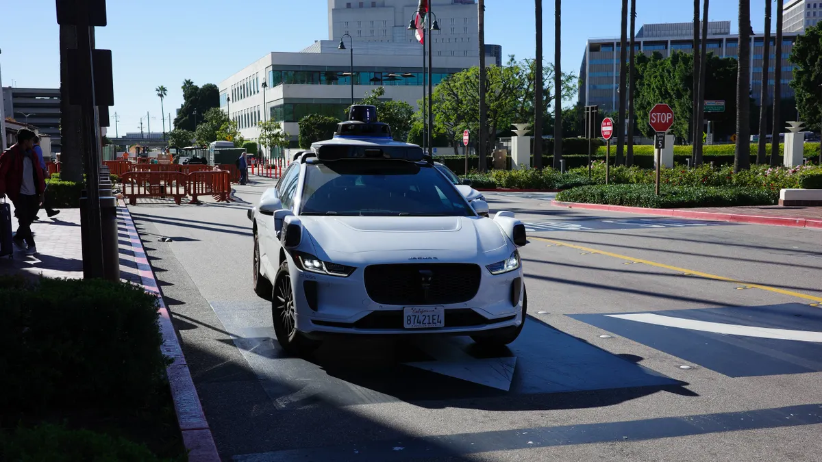 A white vehicle outfitted with many sensors stopped at a curb in with people and buildings in the background.