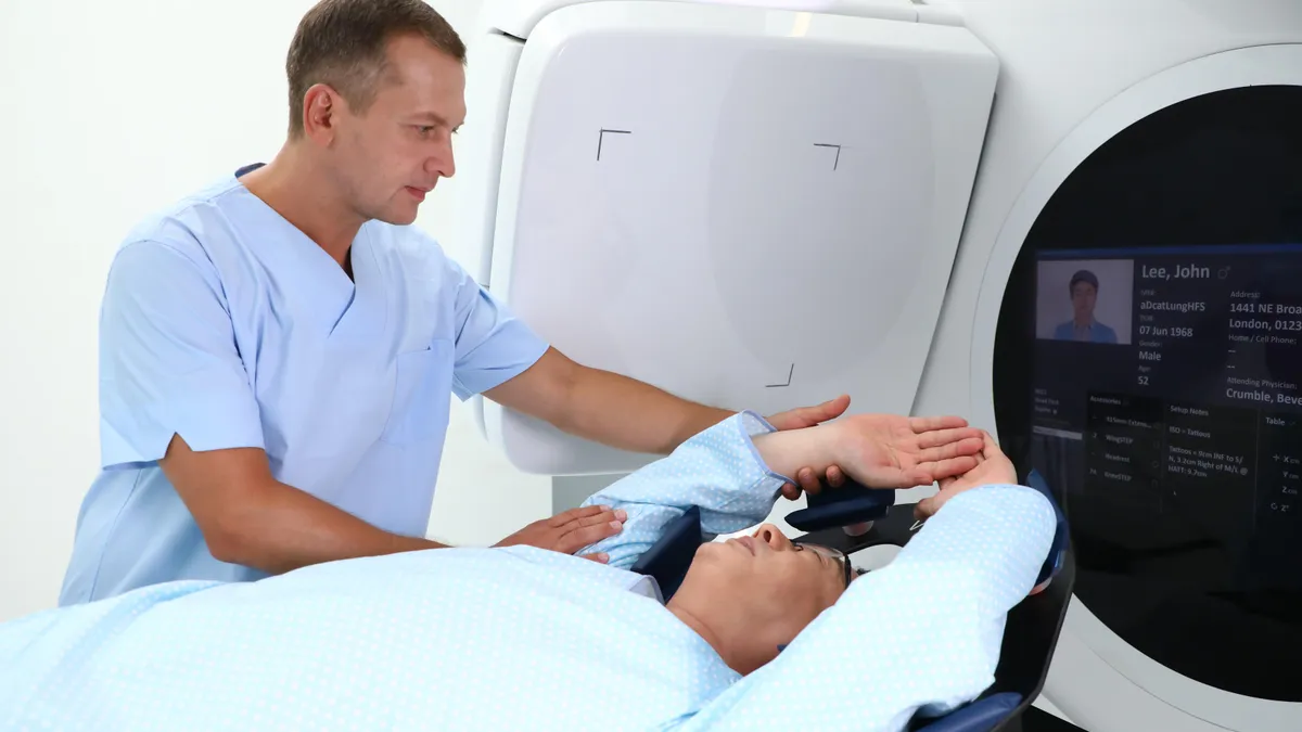 A person lays in front of a radiation therapy machine with both arms up while another person assists them.