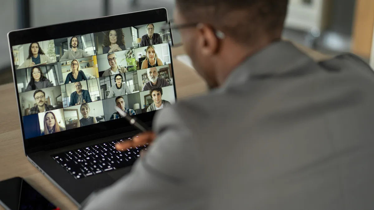 an executive in business attire takes a video call. In the laptop before him there are multiple meeting participants