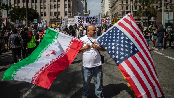A man holds an upside-down American flag and an Iranian flag as he marches down a street. Behind him, other people hold signs, one of which says, "No War On Iran."