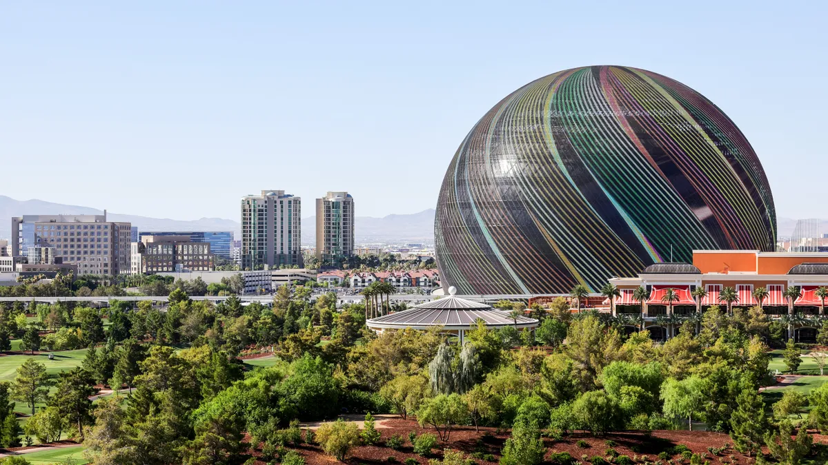 The Sphere and other buildings in the Las Vegas skyline are shown on a clear day, with greenery in the foreground.